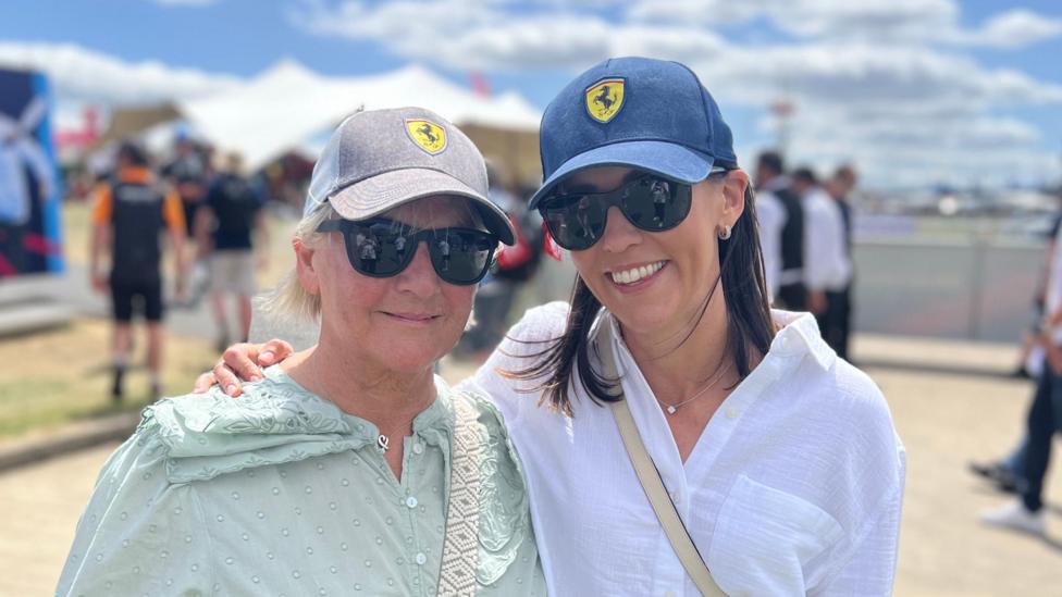 Two women in Ferrari baseball caps stand side-by-side on a bright and sunny day.
