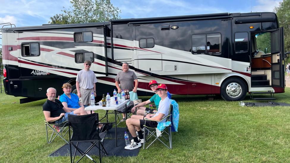 A group of people sat at a picnic table beside a large RV that is parked on a field.