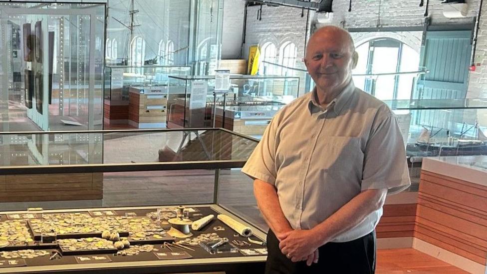 Two men, one in his late 30s on the left and the other inhis late 60s on the right, stood in front of a display of treasure, including several historical gold coins from the reign of Ferdinand and Isabella of Spain, at the National Waterfront Museum in Swansea.