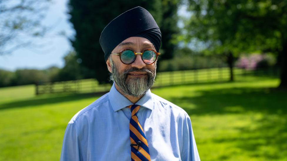 A man stands in his garden, smiling and looking at the camera, wearing a turban and the striped tie of his great grandfather's War regiment
