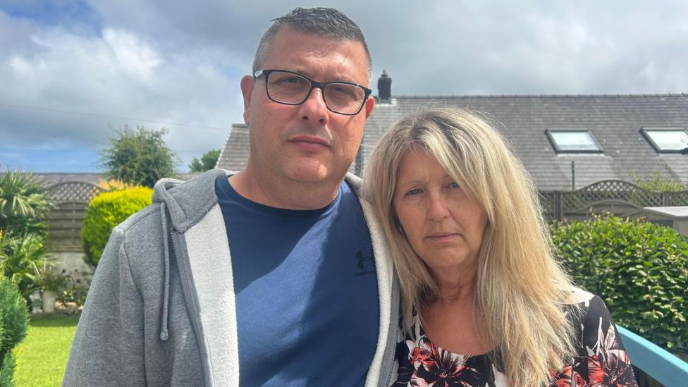 A man and a woman looking at the camera. They are stood in the garden. It is a head and shoulders shot. The man has short greying hair, black-rimmed glasses and a blue t-shirt under a grey woolly hoody. The woman has shoulder-length blonde hair and is wearing a black, white and orange floral print dress.