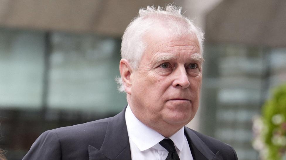 Close-up portrait of a person with short white hair wearing a dark suit jacket, white shirt and black tie. The person is positioned in the foreground and facing slightly to the side. The background is out of focus, showing glass windows, a light-coloured building structure and greenery. No bags, bins, signs or markers are visible.