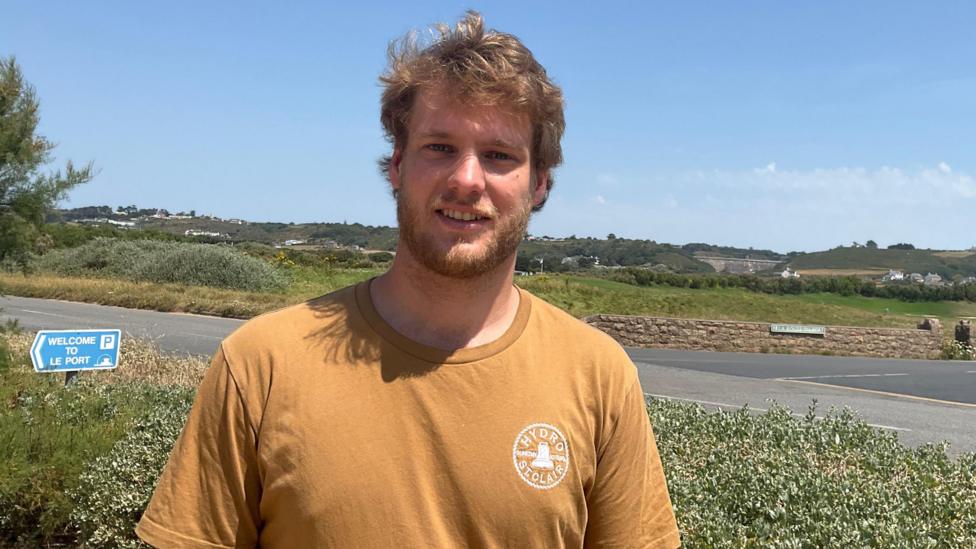 Matt Topman pictured with Les Mielles visible in the background. He is wearing a mustard yellow tshirt, has blonde wavy hair and a beard. He is smiling. A blue sky is pictured above him. 