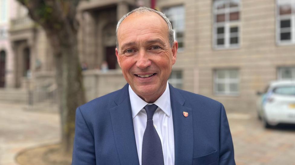 Chief Minister Lyndon Farnham pictured in a suit and tie in the Royal Square. He is smiling. The states building is visible behind him.