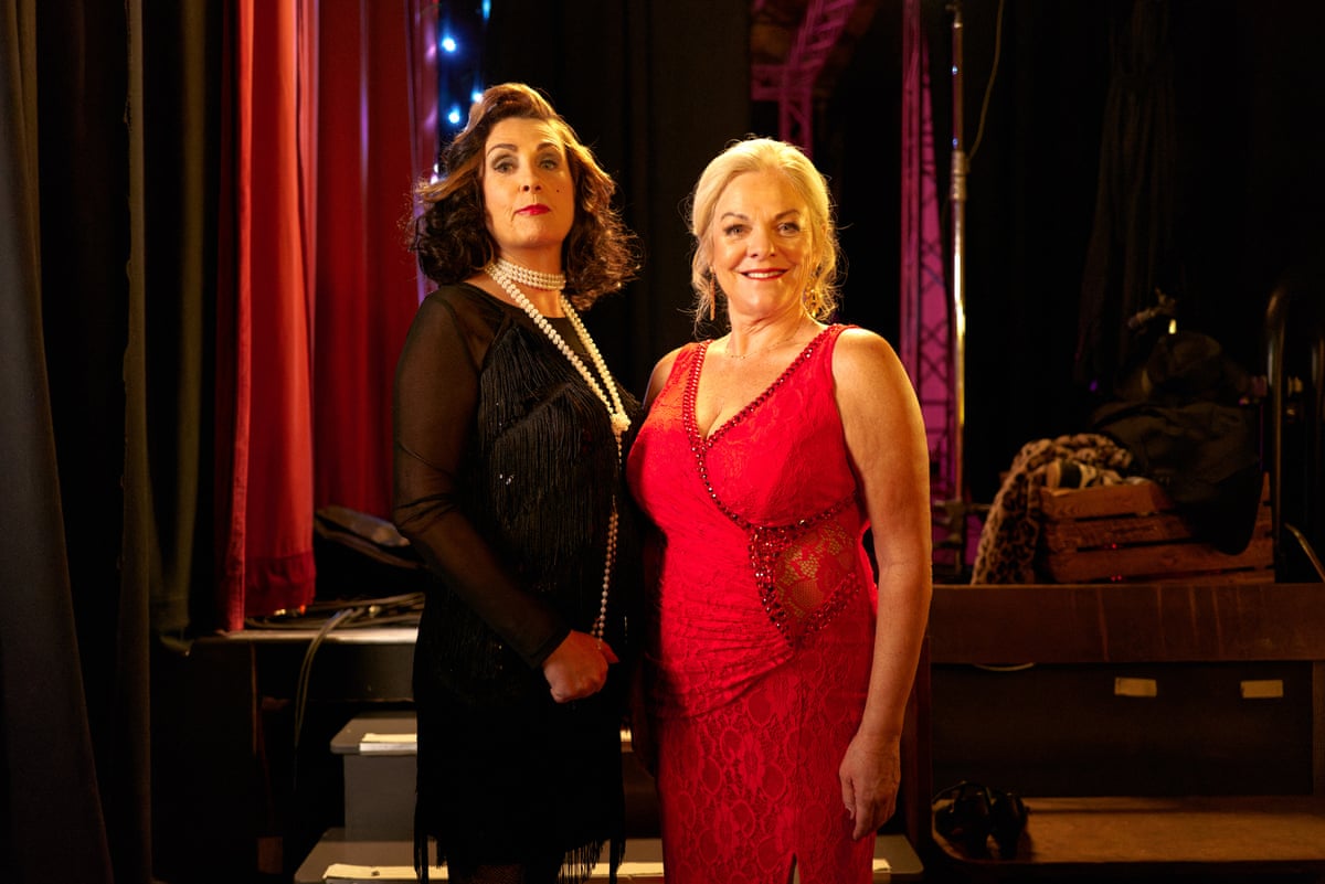 A tall dark woman in black and a blond woman in red evening dresses with stage curtains behind