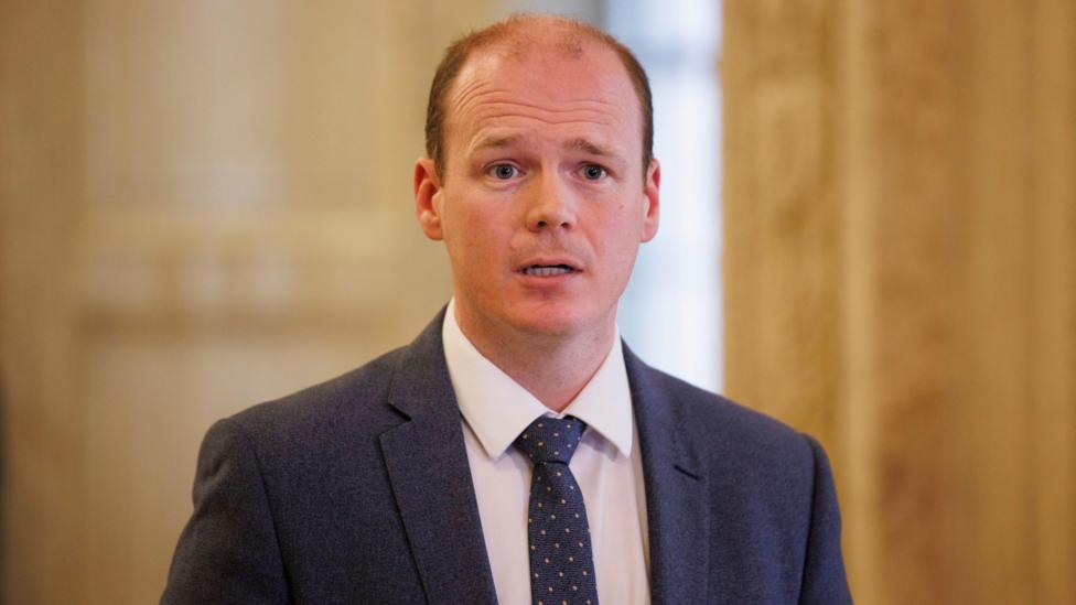 Communities Minister Gordon Lyons, with balding brown hair, wearing a white shirt, navy tie and navy suit.