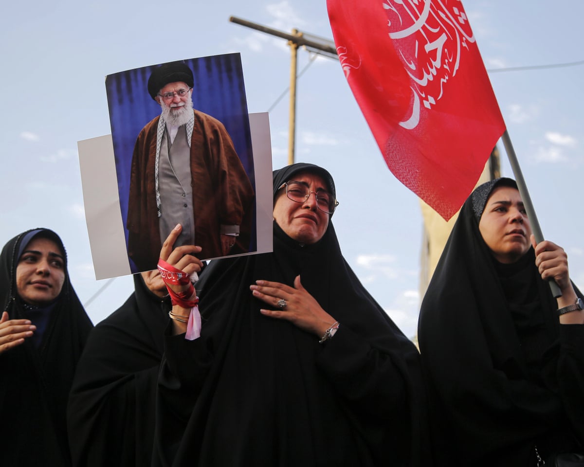 Three women wearing black hold a flag and a photo of former supreme leader Ali Khamenei as part of a crowd of mourners.