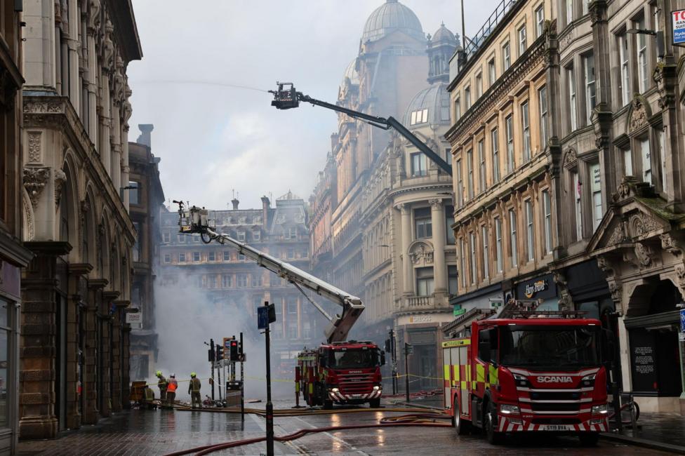 Fire engines at the site of the Union Corner fire in Glasgow. Firefighters use two large hydraulic lifts to aim water from hoses into the smoking building. It is a built up area with a street lined with buildings several floors high.