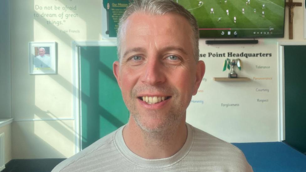 Paddy McMahon, head of St Patrick's RC Primary School, who has short grey haor and a grey top stands in a room in the school with football on a TV screen behind him.