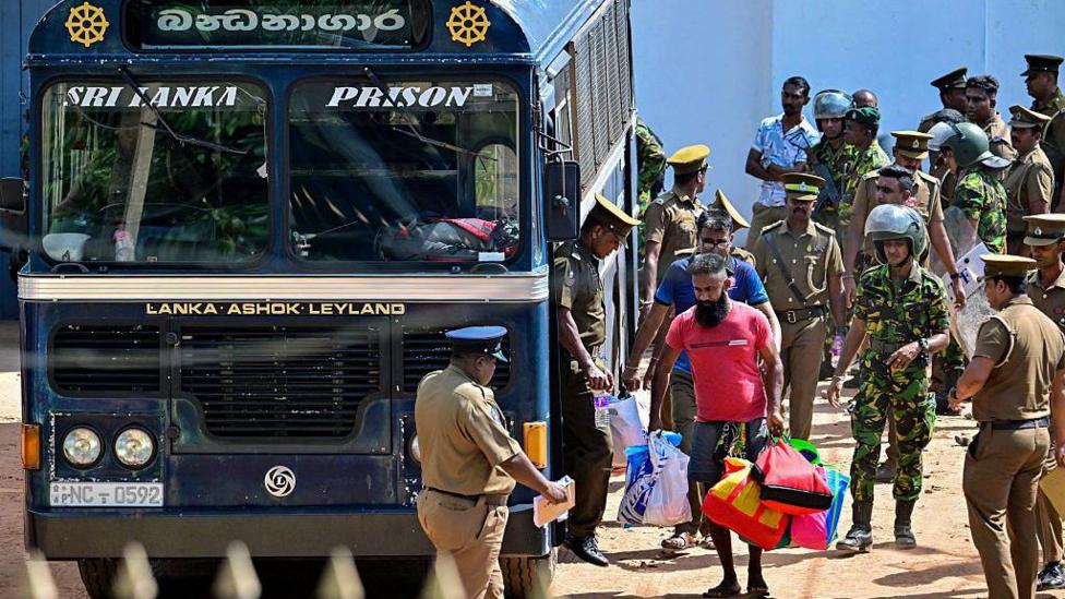 Prisoners being escorted to a blue bus which bears the words Sri Lanka Prison - one man wears a red T-shirt and carries bags. Military officers can be seen around the bus 