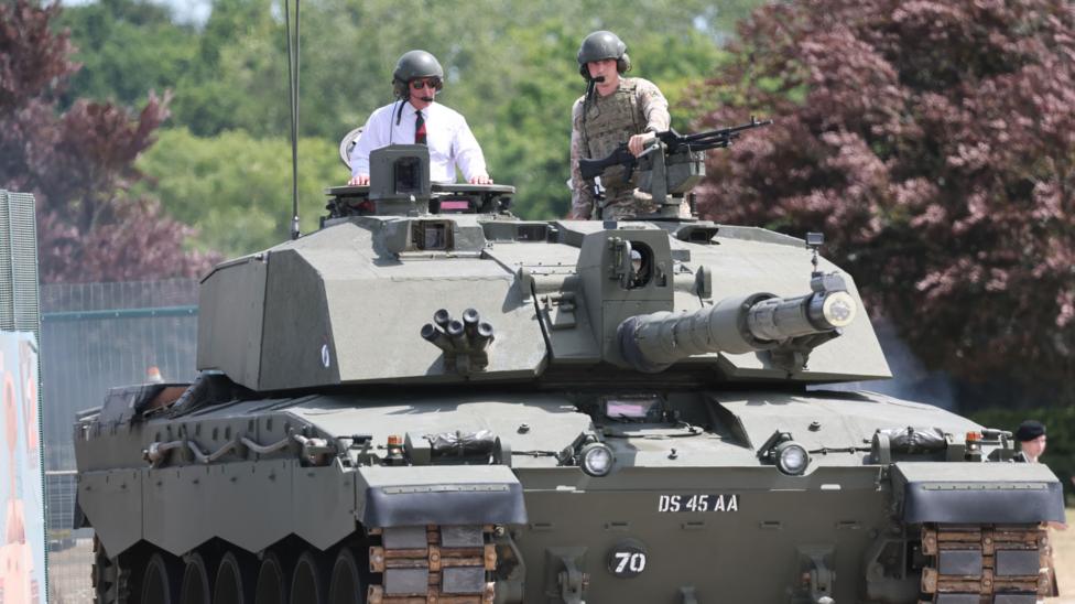 King Charles has the top half of his body sticking out of the top of a tank, he is wearing a white shirt with a red and green tie and some sunglasses. He is also wearing a green helmet with ear defenders and a microphone. Beside him is a man in camouflaged military uniform who is holding a gun and wearing the same style of helmet, ear defenders and microphone as the King.