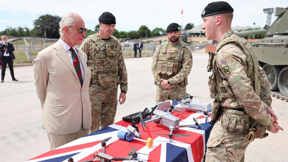 King Charles in a cream coloured suit with a red and green tie and sunglasses. He is talking to three men dressed in camouflaged military uniform, with a table full of drones, covered in a British flag, in between them. There is a tank in the background.