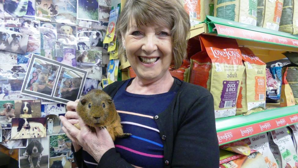Barbara is holding a guinea pig in her pet shop with bags of food on shelves behind her. She is smiling.