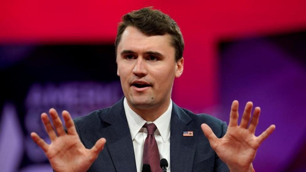 A man gestures with his hands as he talks in front of a red and purple background