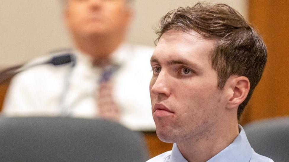 A man with short wavy brown hair and beard stubble looks forward almost expressionless