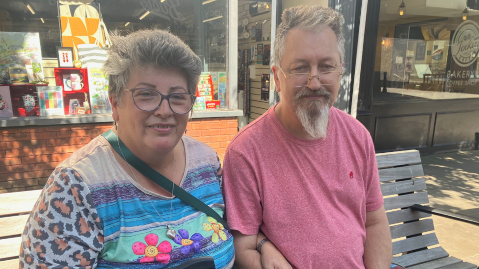 Two people sat on a bench staring at the camera. A man in a pink shirt, glasses and beard, and woman with grey hair and glasses 