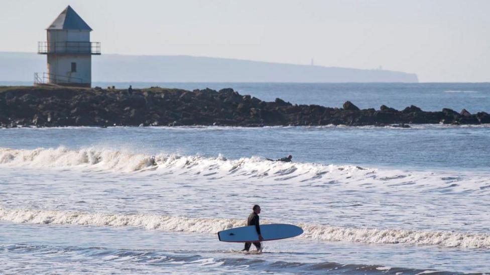 A surfer walking into the sea at Porthcawl on a sunny day