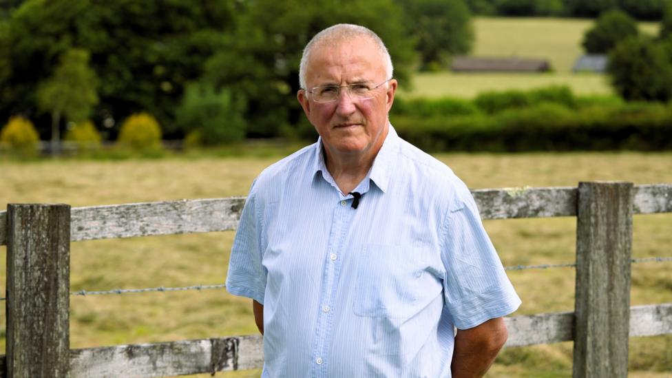 Rhodri Williams, with grey hair and glasses, wears a short sleeved blue shirt. He is standing in front of a wooden fence with a field and trees blurred in the background.