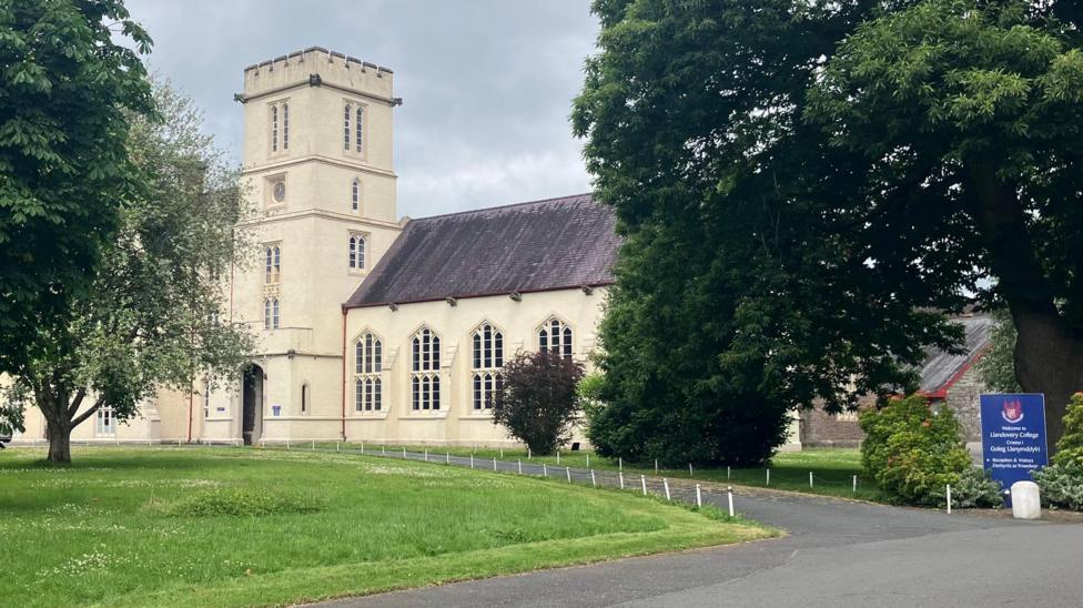 An old cream stone building in grounds with a blue sign saying Welcome to Llandovery College in English and in Welsh at the end of the drive. 