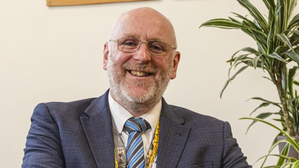 A man with a beard and gold-rimmed glasses wearing a blue checked jacket, a striped tie and lanyard
