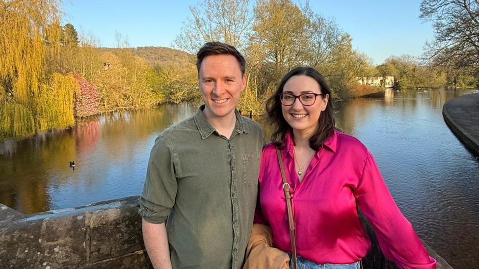 Laura-May Nardella and her husband Ryan Monk are smiling at the camera, with a lake and trees behind them. Laura-May is wearing a bright pink shirt with glasses and has brown shoulder-length hair. Ryan is wearing a great shirt and has short brown hair