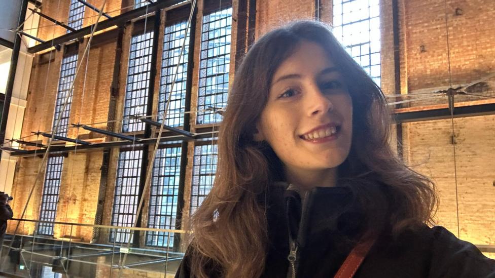 Emma is smiling for the camera, taking a selfie. She has long brown hair and behind her is a big bricked building with big windows and architectural features