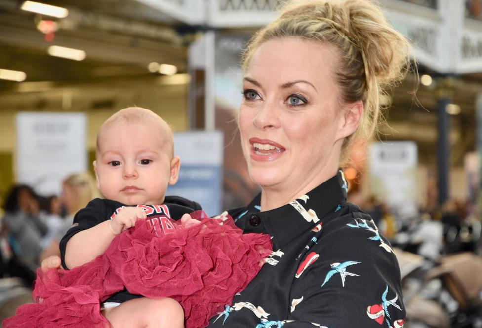 A woman with blonde hair loosely tied in a bun, and wearing a black shirt with birds and hearts printed on it, holds a baby, wearing a black tee and red tutu, close to her chest.