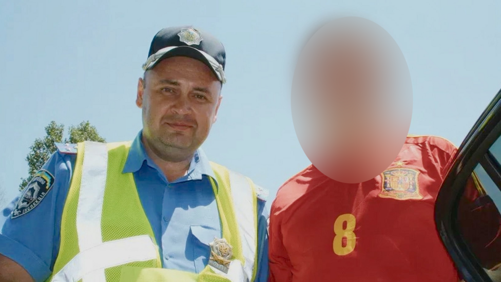 Temerbek is pictured in a black police cap, blue police shirt and yellow hi-vis vest, looking at the camera with a slight smile. Next to him is a figure in a red football shirt, whose face has been blurred.