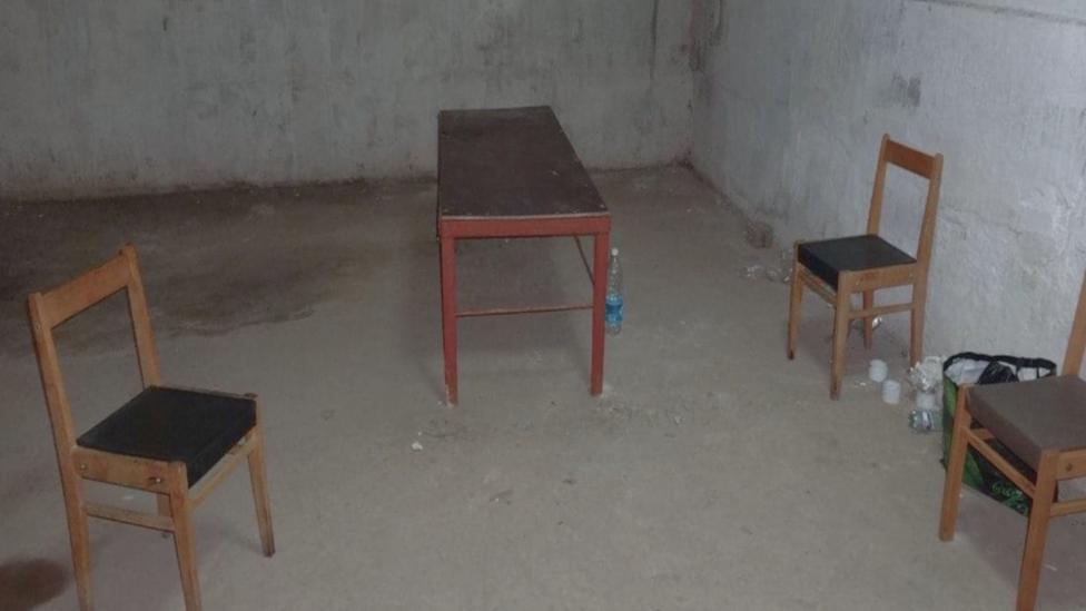 A bare room with concrete walls and floor. In it are three wooden chairs and a small wooden table, with a few plastic water bottles on the floor.