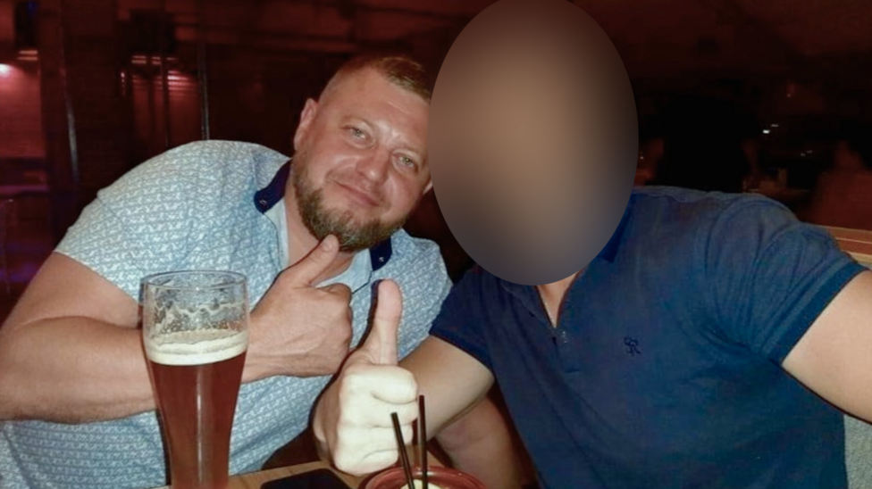 Two men with large glasses in front of them giving a thumbs up sign to the camera. The one on the left, Ruslan Yeriomichev, has a trimmed beard and close-cropped hair, and is wearing a light patterned T-shirt. The face of the man on the right has been blurred.
