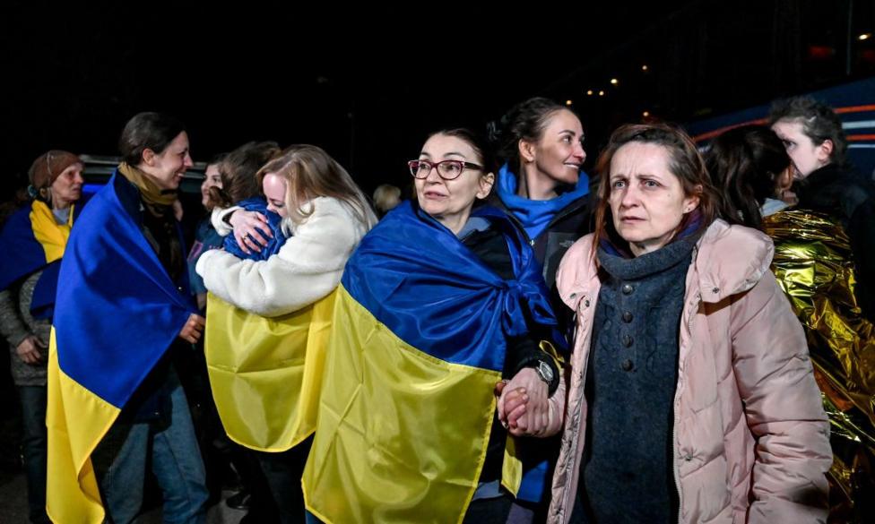 Several women wrapped in blue and yellow Ukrainian flags. Two women are hugging the background, others are smiling. Liudmyla is looking to her right while holding the hand of a woman in a pink coat on her left.