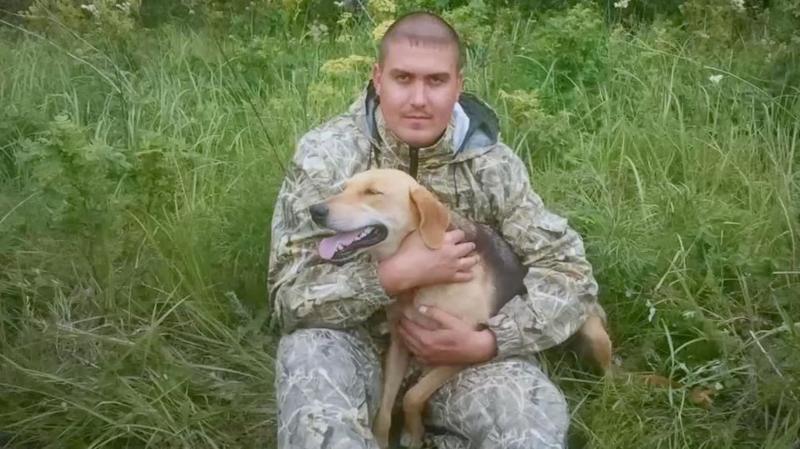 Andrey Spivak sitting in long grass holding a beige and black dog. He is wearing a camouflage-style outdoor suit, has hair shaved closed to his head and is looking directly at the camera.