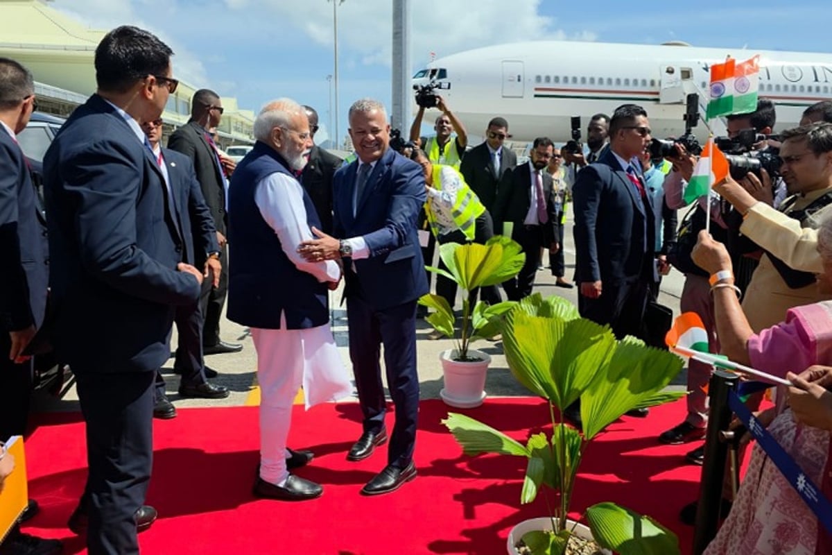 Modi stands on a red carpet at the airport with a plane in the background as officials stand by and people wave Indian flags.