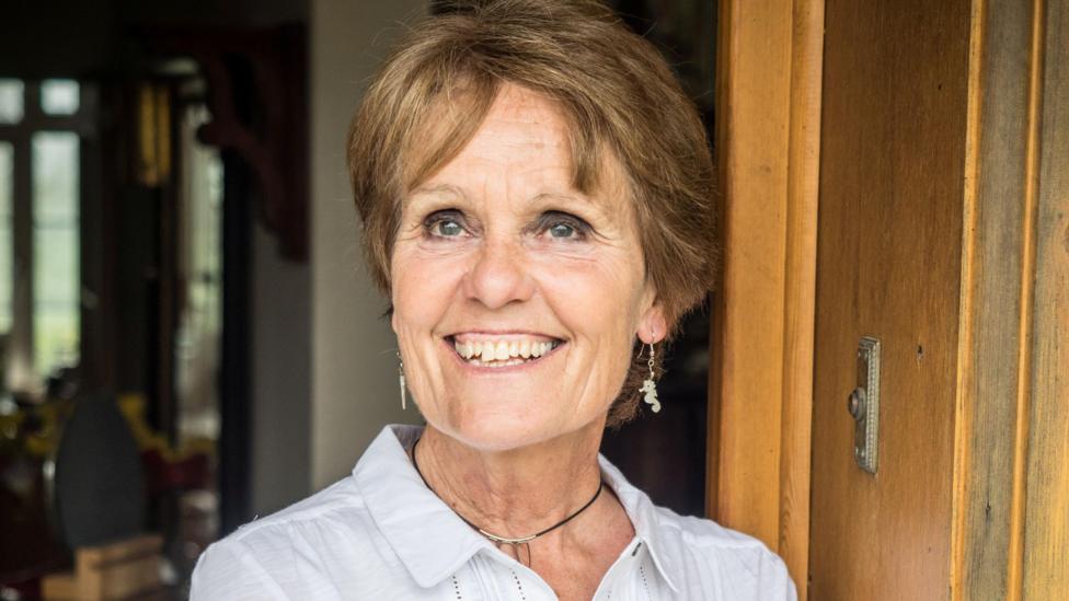 Gina Rippon. She has short brown hair and blue eyes. She is wearing a white linen shirt and black necklace, paired with seahorse shaped pendant earrings. She is leaning on a wooden door. She is smiling and looking at something out of shot.