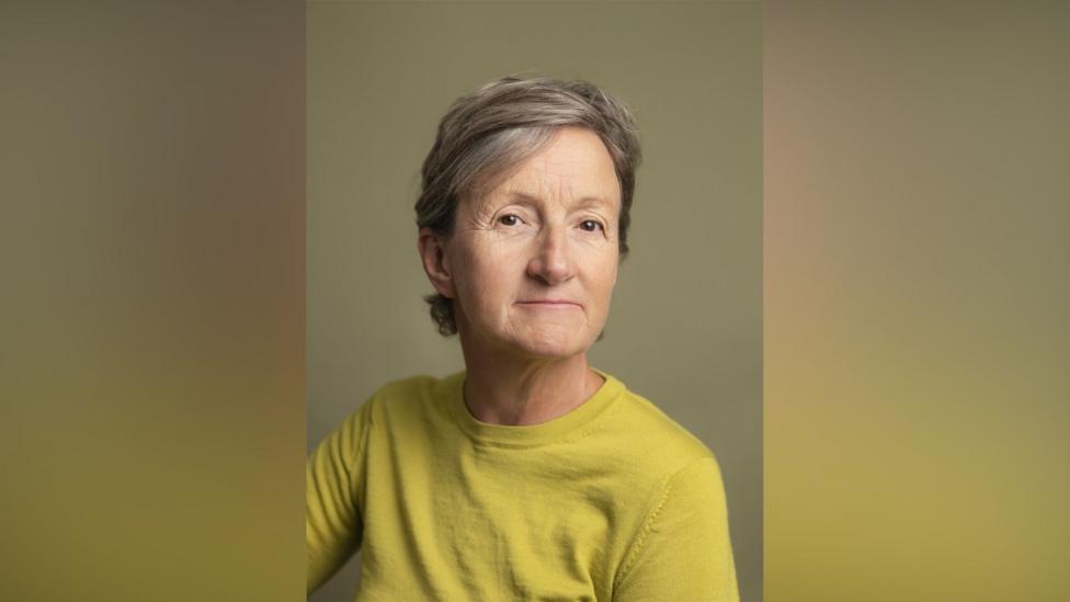Sarah Hendrickx. She has short grey hair and brown eyes. She is wearing an acid green jumper and looking at the camera with a slight smile.