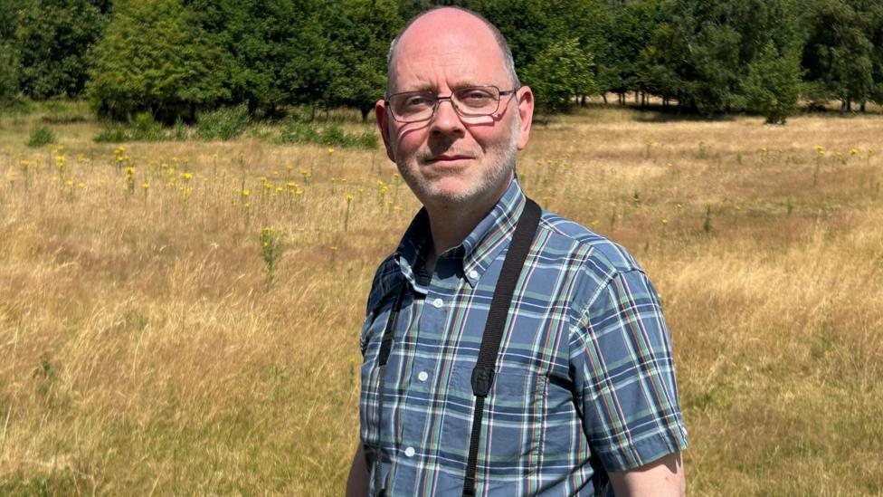 A man, blad on top but with grey hair on the sides of his head, standing in a grassland field with trees in the background. It is clearly a summer's day. He is wearing thin-rimmed glasses and a short-sleeved check shirt and has something out of frame hanging around his neck.