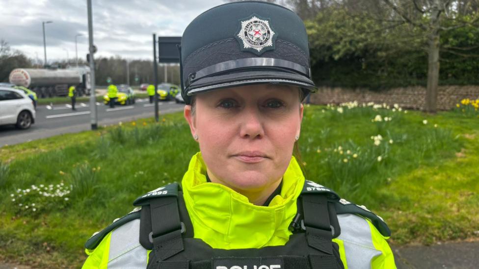 A woman wearing a PSNI uniform looks at the camera. She is wearing a dark-green hat and a fluorescent yellow jacket. Behind her is a patch of grass, and further in the distance is a road on which several other police officers are standing.