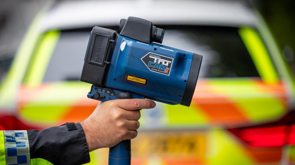A police officer holds a speed detection camera. a police vehicle is in the background