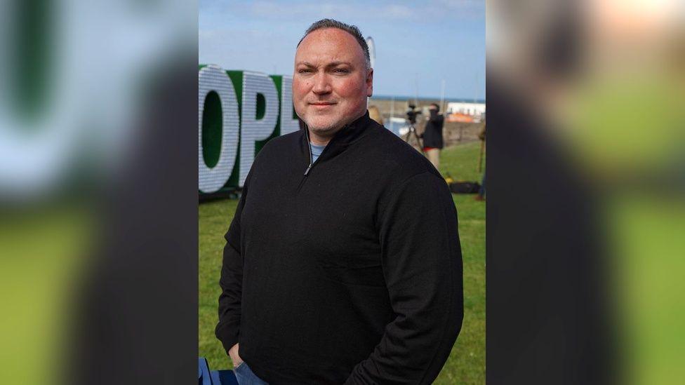 Chris Arthur stands with his hands in his jeans pockets. He is wearing a black top, denim jeans and is stood in front of a sign. The large white letter O and P can be seen over his right shoulder