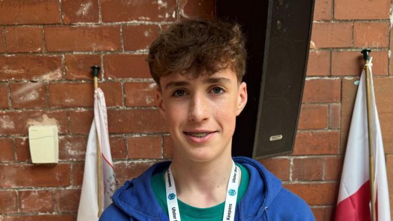 John has short dark hair and is wearing a blue jumper, green top and has a white lanyard around his neck.
