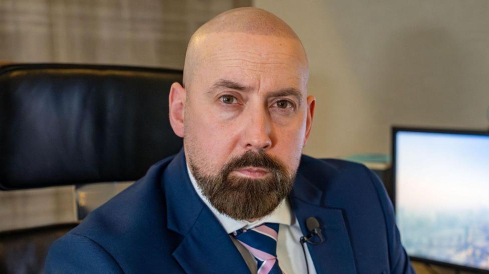 David Kennedy is bald with a brown beard. He wears a navy suit and sits in an office