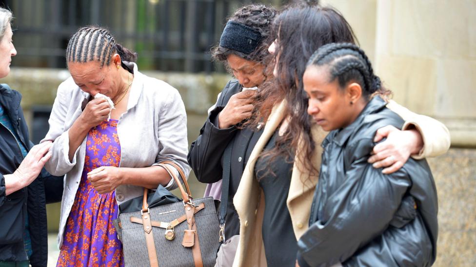 A group of women huddled together outside a court building. Two have their heads bowed and are weeping into handkerchiefs.