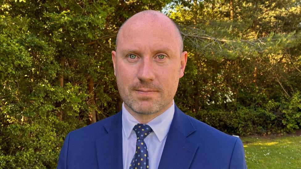 Neil Gray is bald and wears a navy suit. He stands outside in a garden