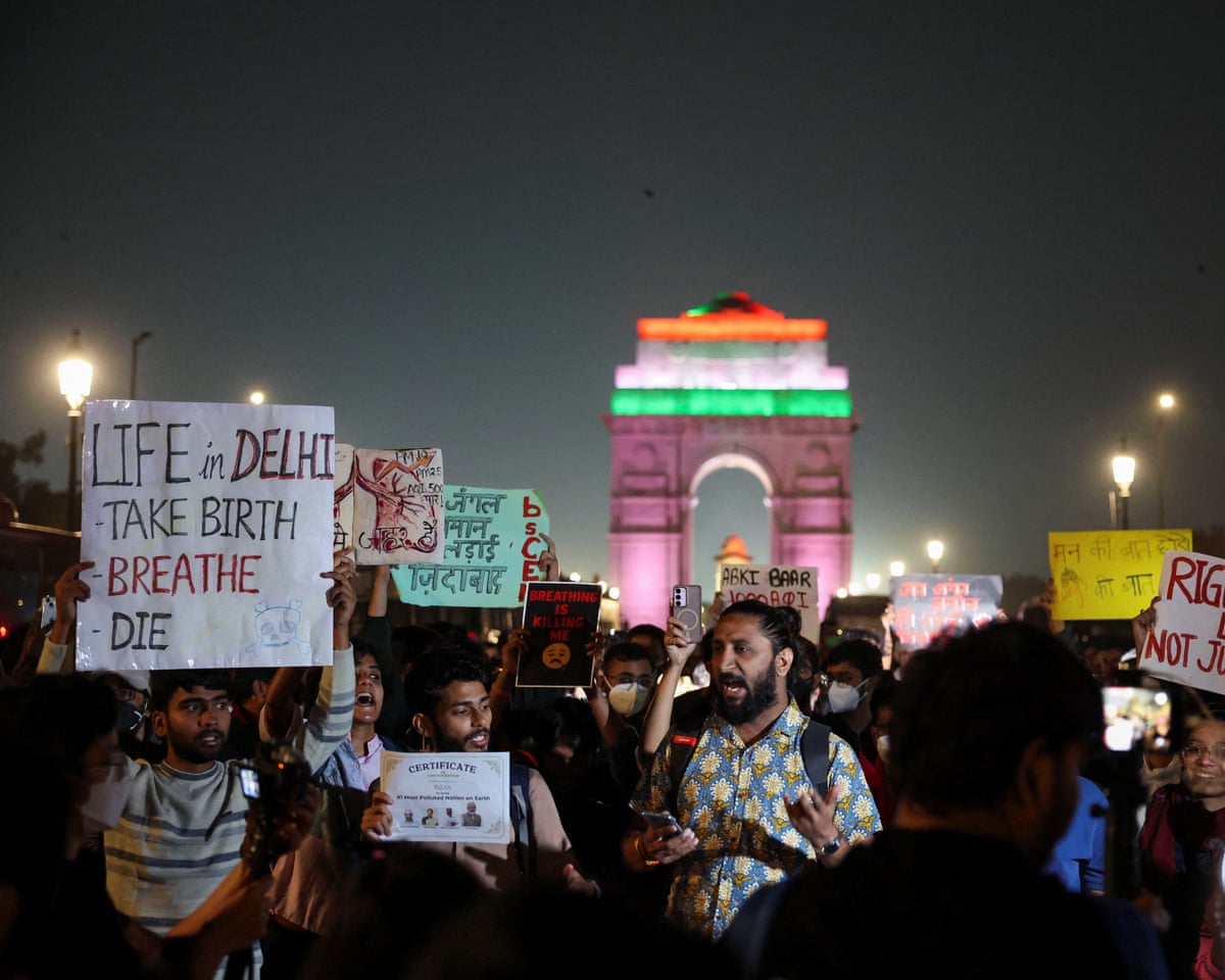 A crowd gathers holding signs that include: ‘Life in Delhi - Take Birth, Breathe, Die’