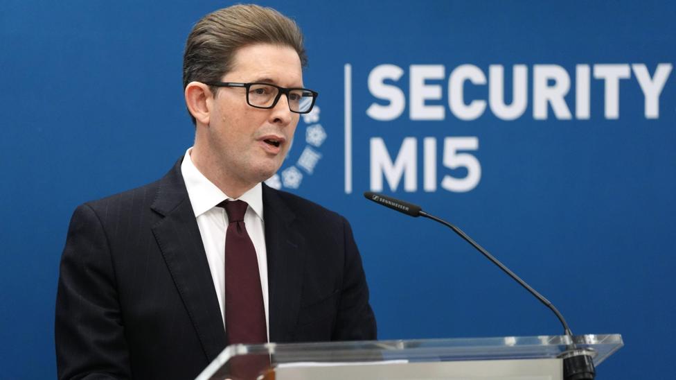 Ken McCallum, Director General of MI5 - a middle-aged man with slicked back dark brown hair, and wearing dark-rimmed rectangular glasses. He is wearing a dark suit, white shirt and navy blue tie.
