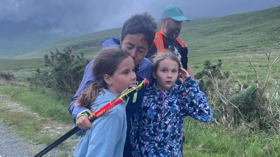 Trish Patterson hugging her twin girls and giving one of them a kiss on the head as she walks along a wild path with a man in high vis in support behind her.