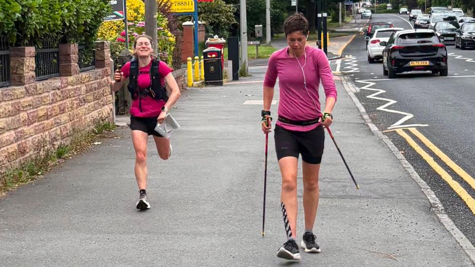 Trish Patterson, wearing a pink top and black shorts and shoes, on the right with walking sticks and walking along a road with cars travelling along. She has headphone in and is focused on the floor. She has tap on the front of her leg.