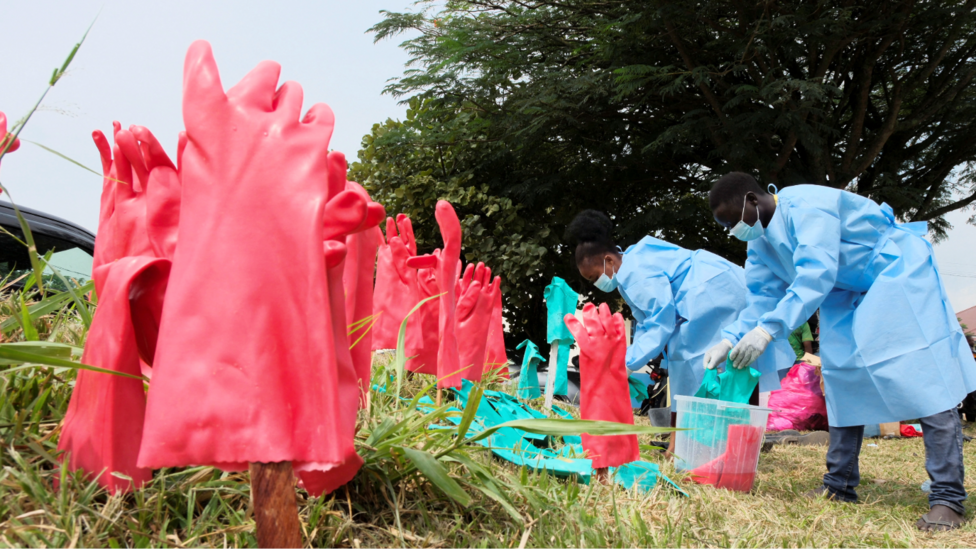 Congolese medical workers in blue personal protective equipment (PPE) sanitise pink and blue rubber gloves in buckets at Ebola treatment centre in Bunia. Gloves already washed dry upright on sticks spoked into the grass.