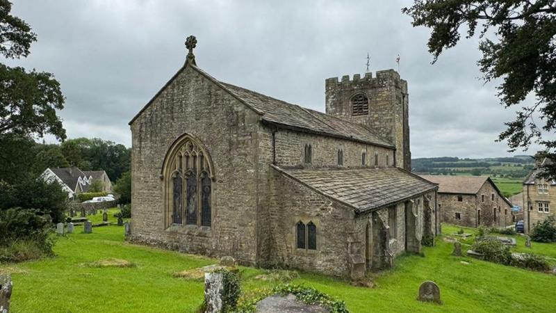 External view of St Wilfrid's Church which is in the village of Melling near Lancaster. Gravestones can be seen in the churchyard with fields in the background.