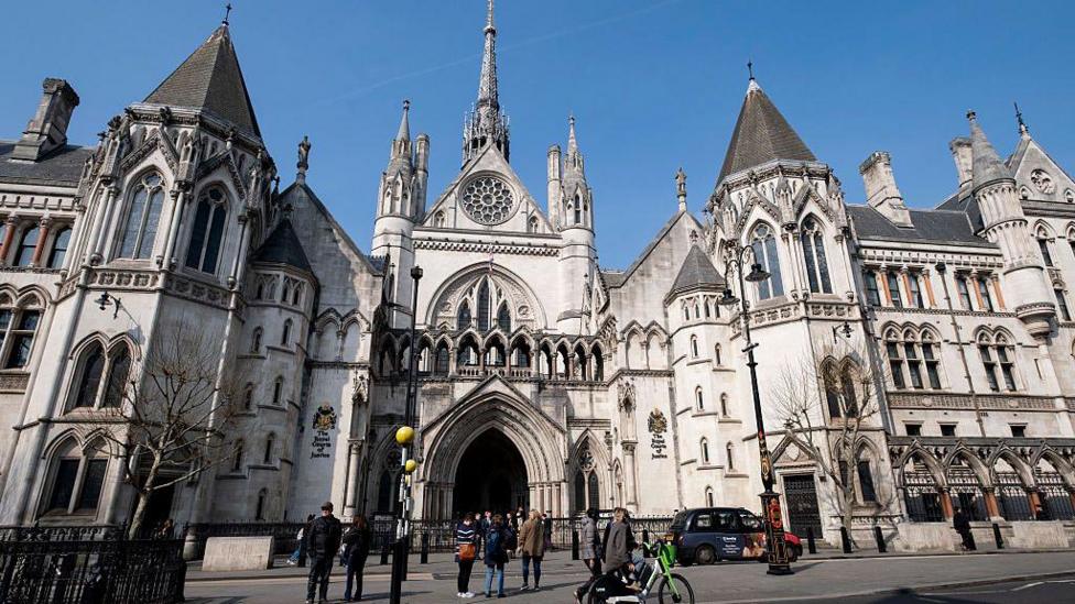 The Royal Courts of Justice against a sunny, clear-blue sky. It is a large grey stone building in the Victorian gothic revival style. A number of people are gathered outside its entrance, and a black taxi cab is driving on the road in front of the building.